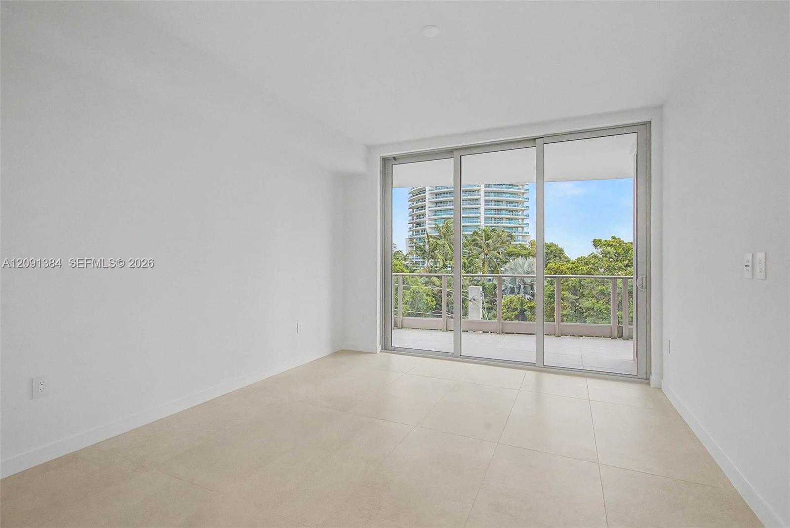 Vacant condo interior with pale tile floors and sliding doors overlooking a tree-filled balcony and high-rise.
