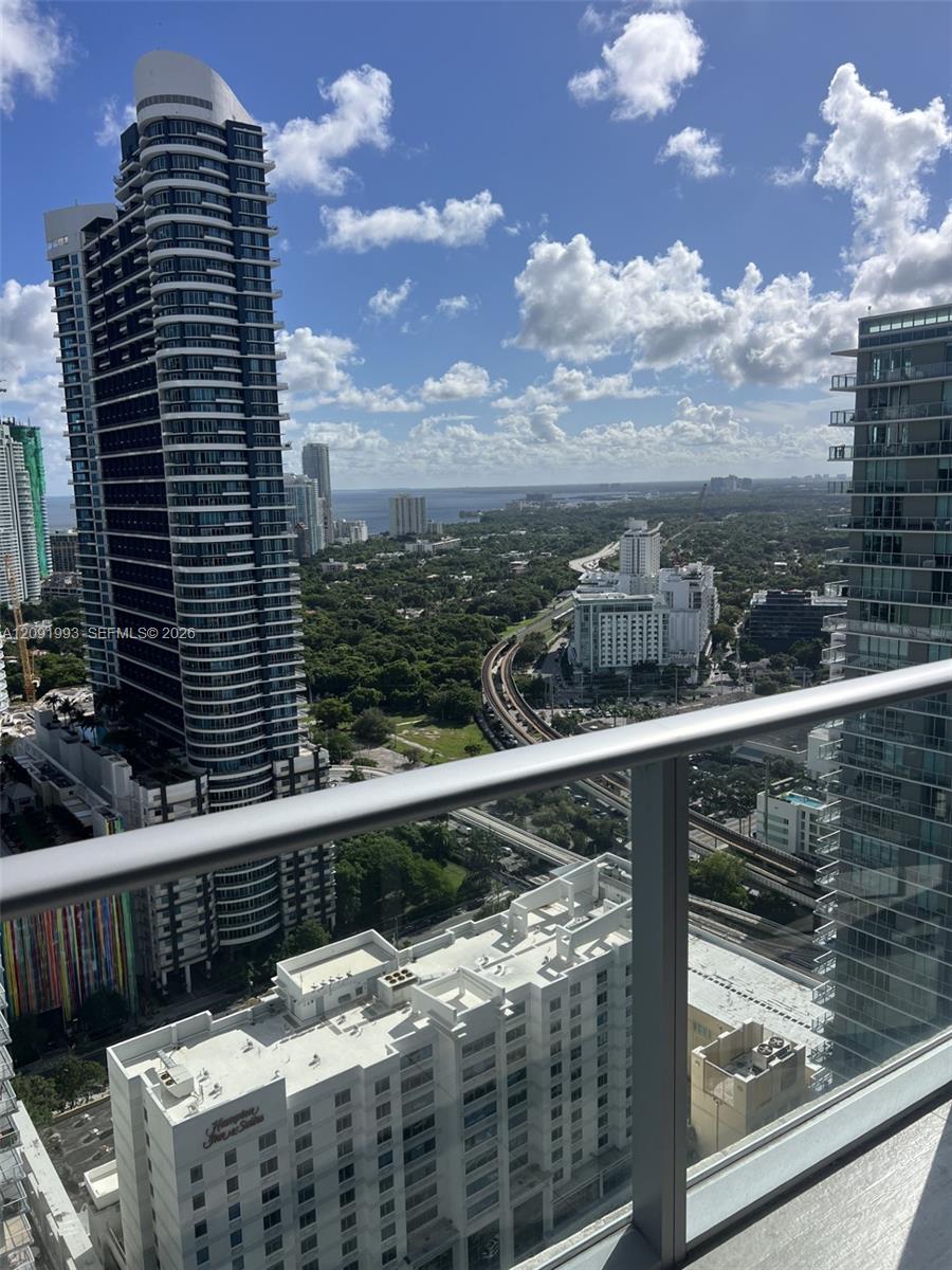 Balcony view over Miami high-rises, transit tracks, green canopy, and water beneath a cloud-filled sky.