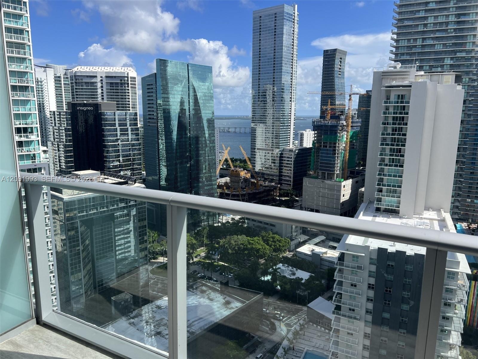 Downtown skyline and waterfront viewed from a glass-railed balcony, with a bridge and construction cranes in the distance.