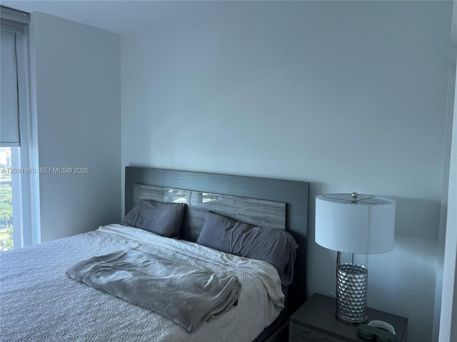 Bedroom with white bedding, a dark headboard, and a silver bedside lamp beside a window.