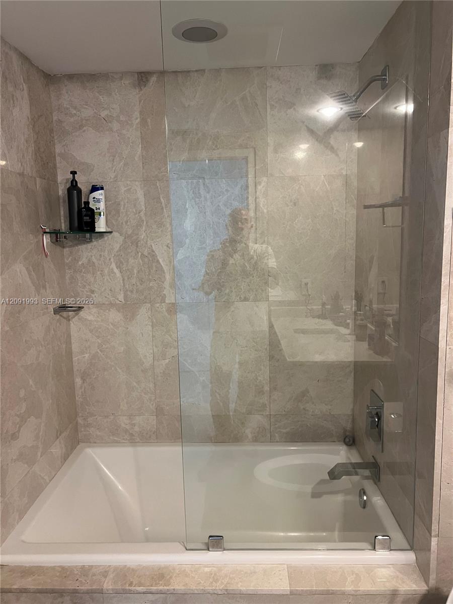 Bathroom bathtub with clear glass screen, beige marble-look tile, and wall-mounted showerhead.