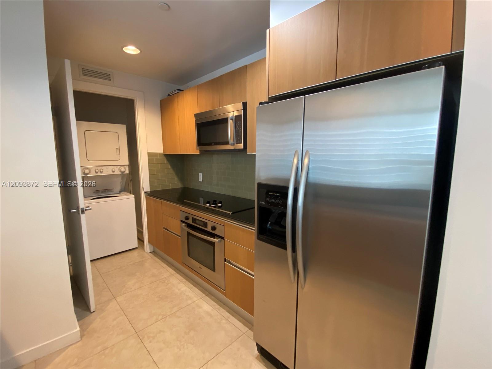 Compact kitchen with light wood cabinets, stainless steel appliances, and a stacked washer and dryer through the doorway.