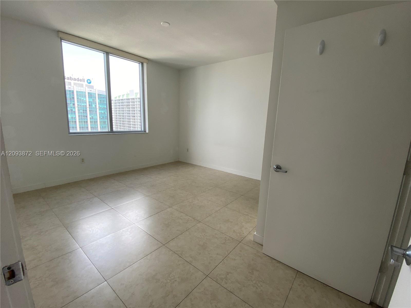 Empty condo room with beige tile floors and a large window overlooking nearby high-rises.