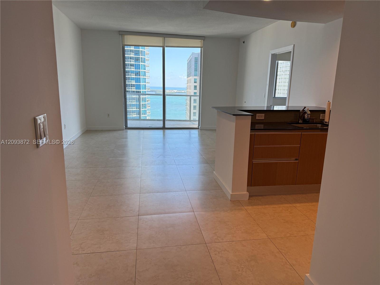 Unfurnished condo living area with tiled floors, a kitchen peninsula, and water views through sliding glass doors.