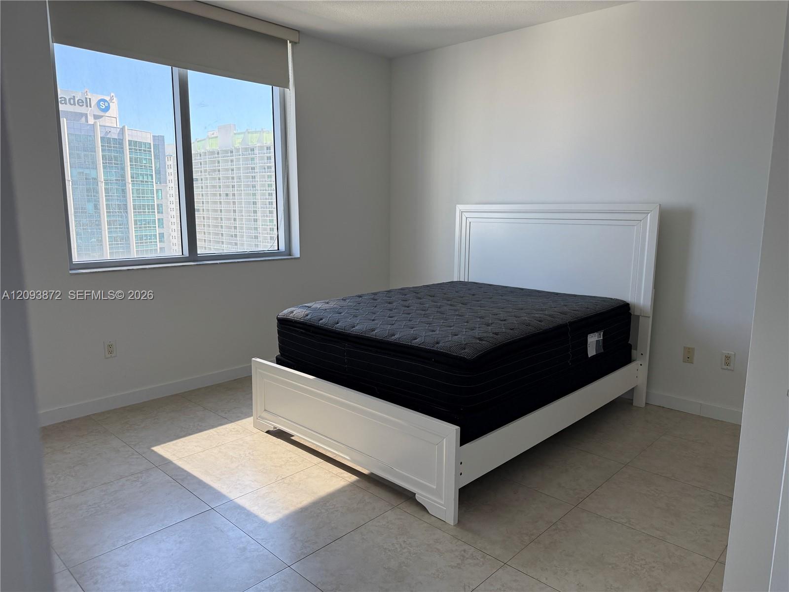Bedroom with a white bed frame and dark mattress beside a large window overlooking neighboring buildings.