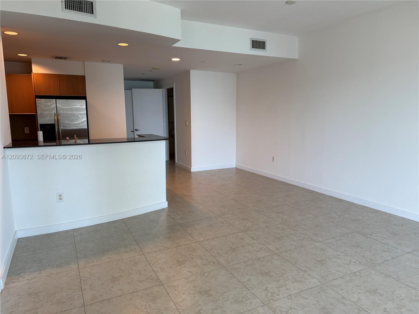 Empty condo living area with tiled floors and an open kitchen with wood cabinets and stainless steel refrigerator.