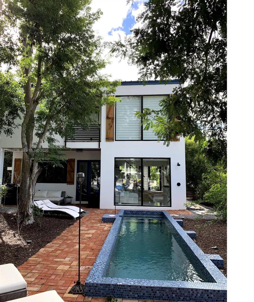 Modern white house with a narrow blue-tiled pool and brick patio, framed by leafy trees.