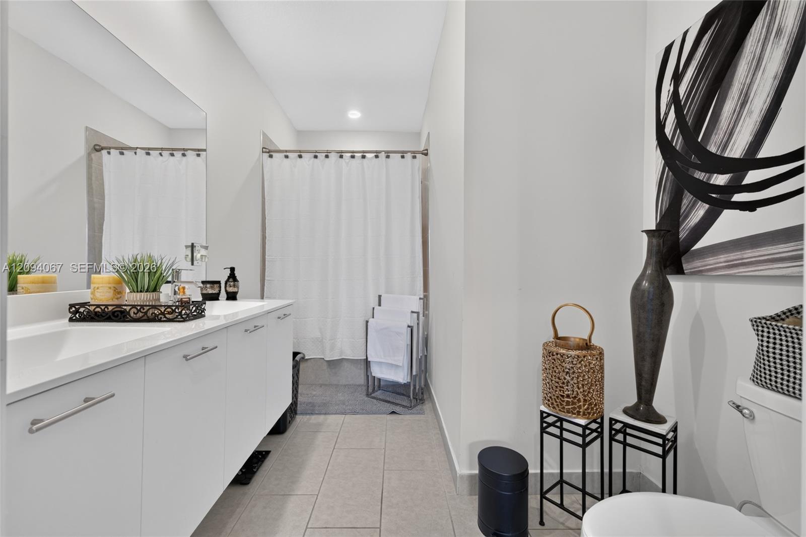 Bathroom with a long white vanity, tiled floor, and shower curtain, accented by abstract art and decorative vases.