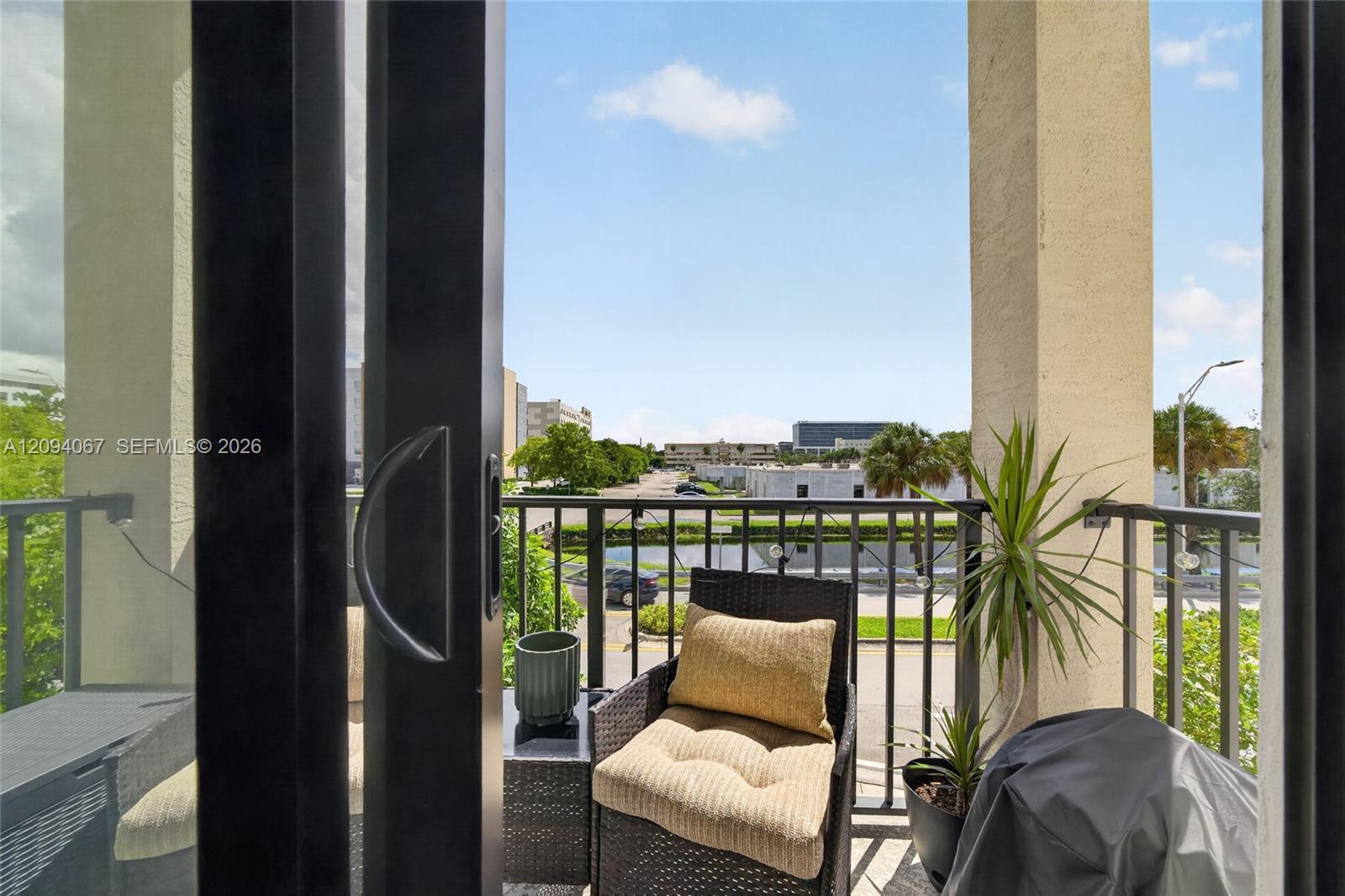 Apartment balcony with wicker seating, potted plants, and a view of nearby buildings.