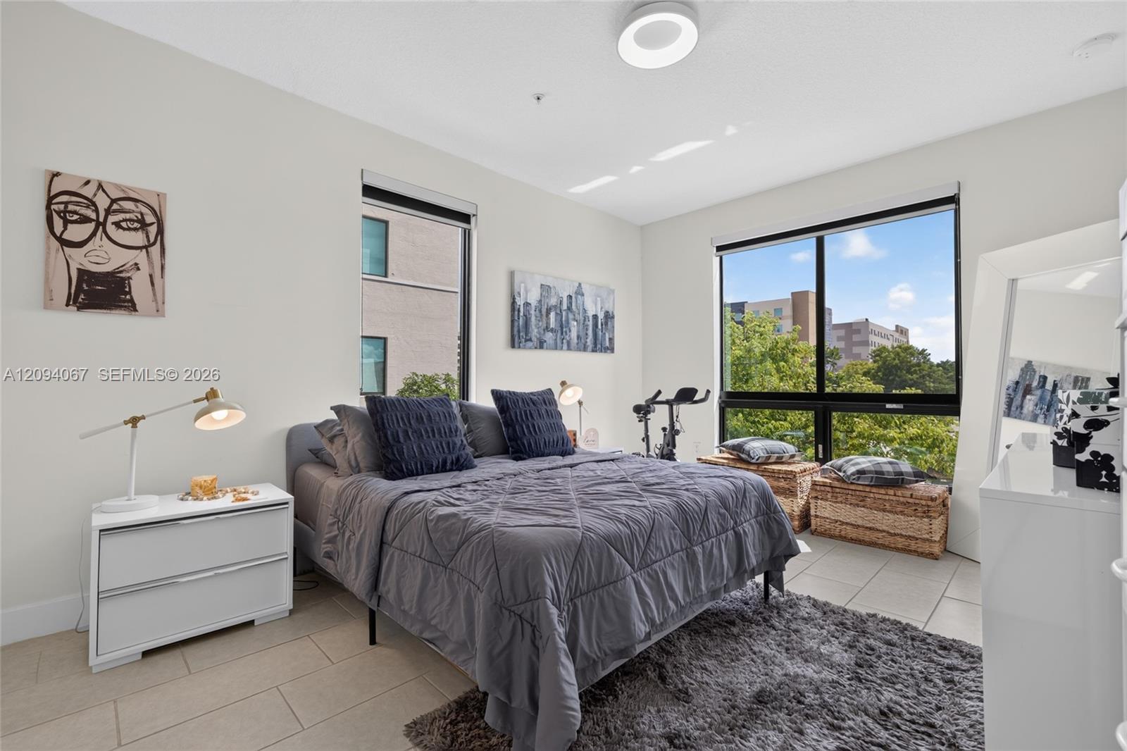 Furnished bedroom with a gray bed, tiled floor, and large windows overlooking trees.