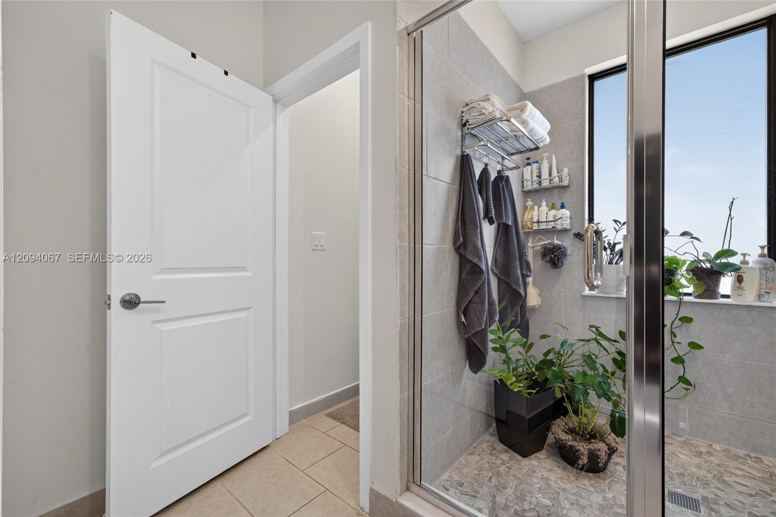 Bathroom with a glass-enclosed tiled shower, towels, and potted plants beside a large window.