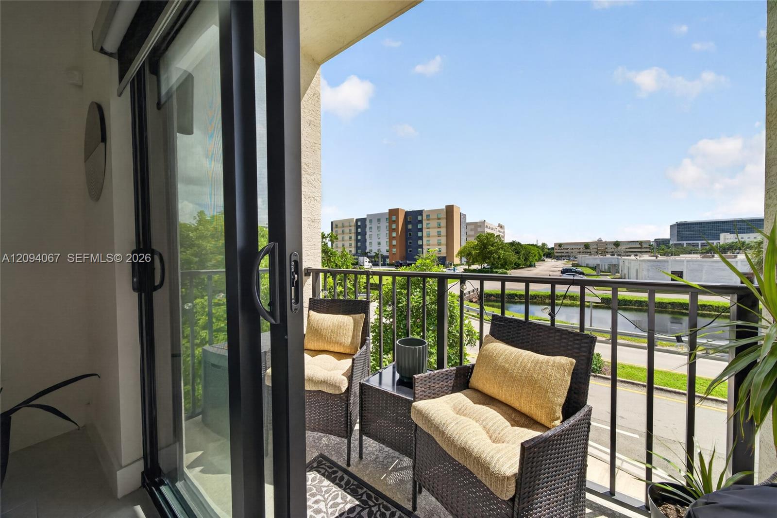 Furnished balcony with wicker chairs overlooking a canal, road, and nearby buildings.