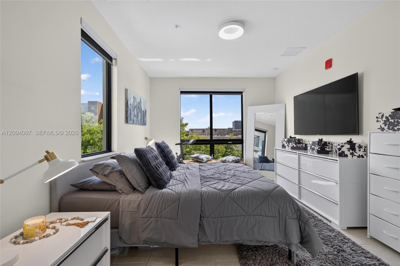 Staged bedroom with a gray bed, white dressers, and large windows overlooking trees and nearby buildings.