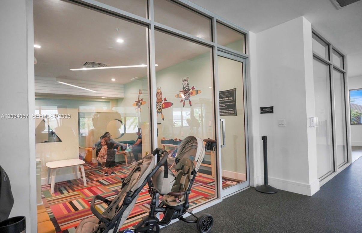 Glass-walled children's playroom with a colorful rug and strollers in the hallway.