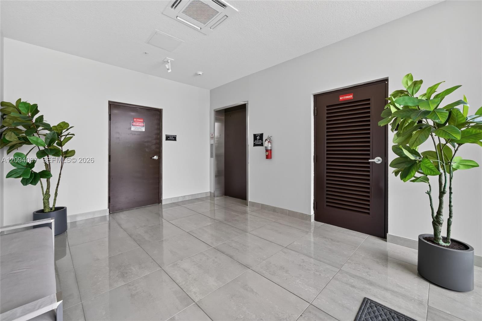 Tiled building hallway with an elevator, dark doors, and potted plants.