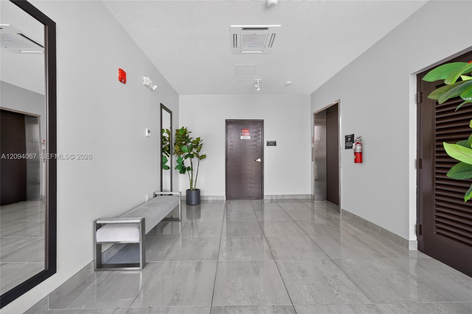 Modern building lobby with glossy gray tile floors, elevator doors, a bench, and a potted plant.