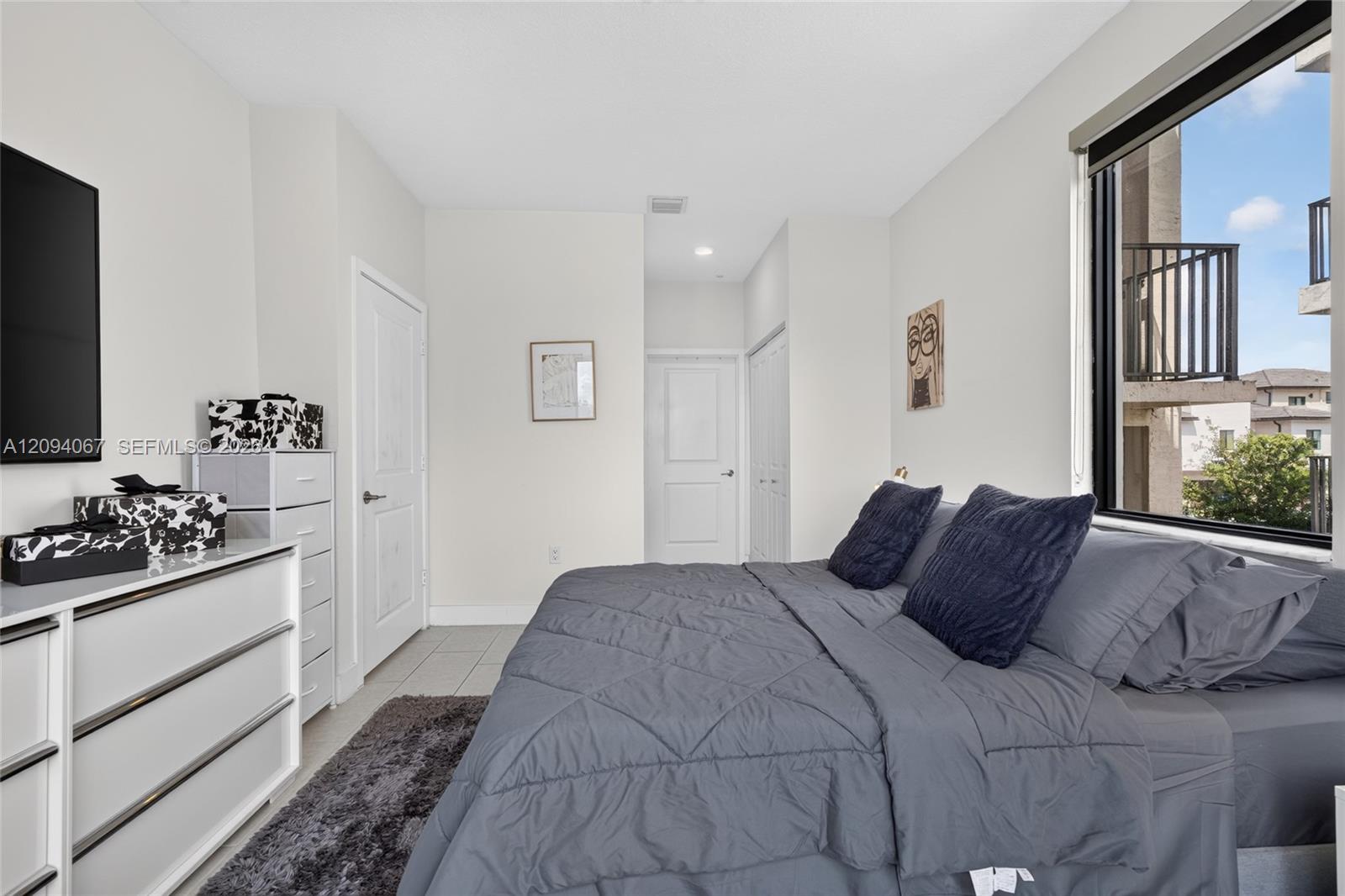 Furnished bedroom with a gray bed and large window overlooking neighboring buildings.