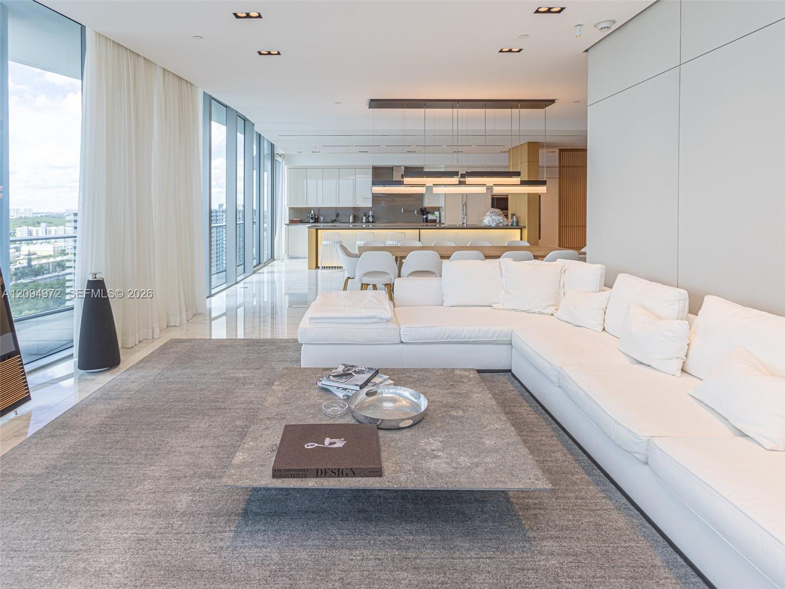 Furnished condo living room with a white sectional, floor-to-ceiling windows, and an open kitchen.