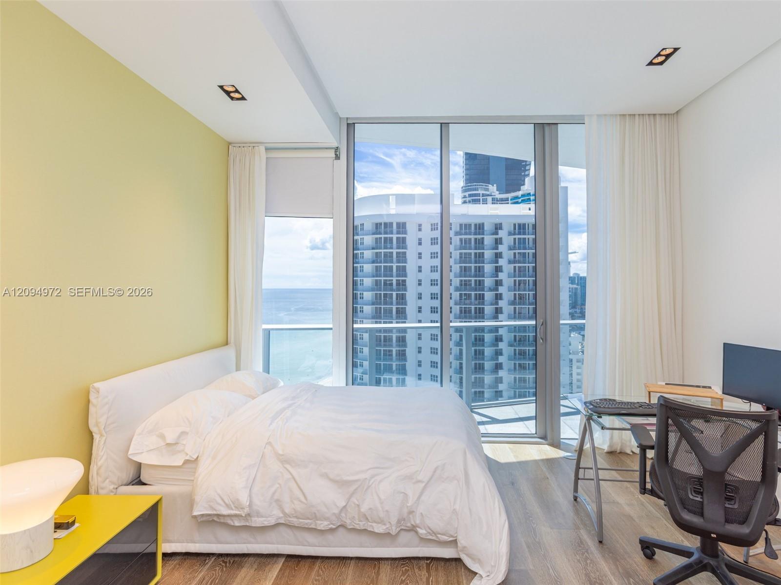 Bedroom with a white bed, yellow accent wall, and floor-to-ceiling windows overlooking the ocean and high-rises.