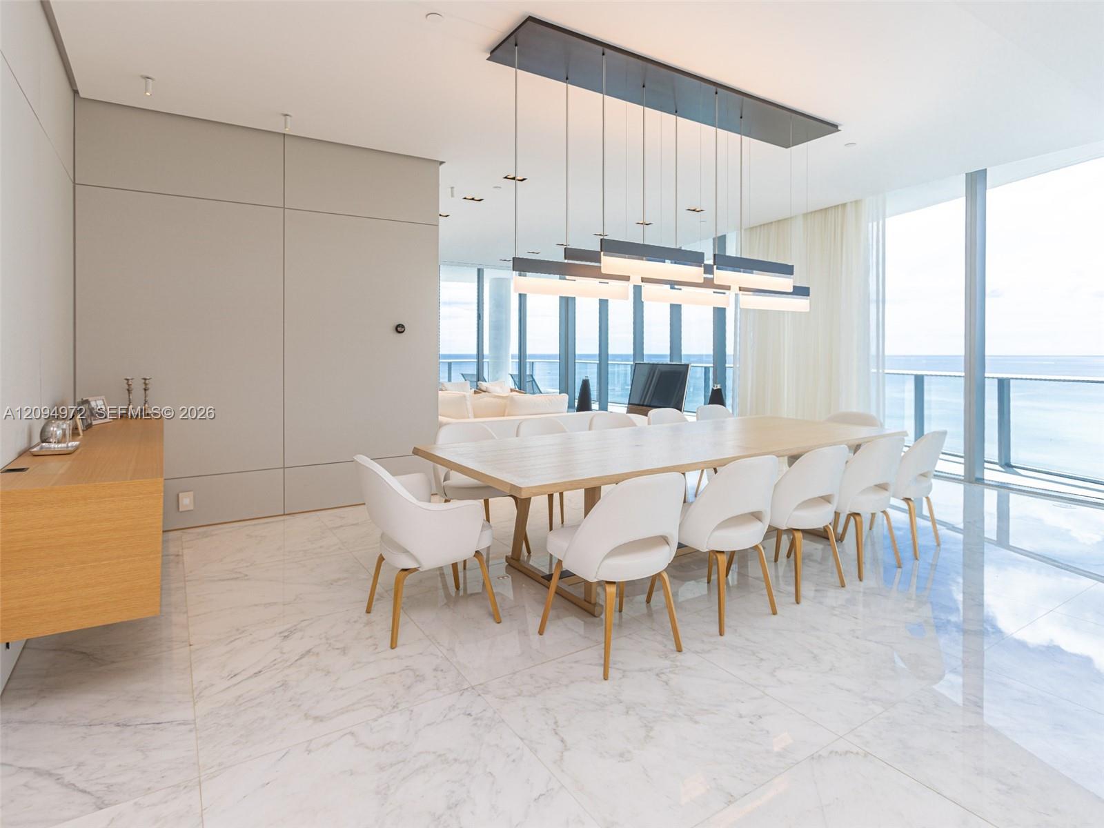 Modern dining area with white chairs, marble floors, and floor-to-ceiling ocean-view windows.