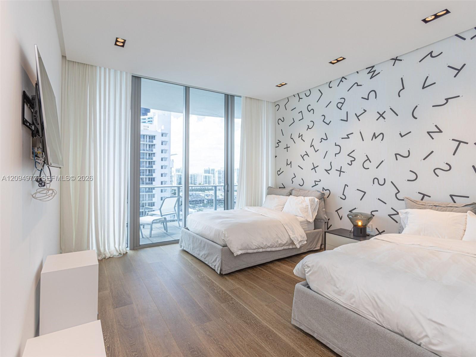 Bedroom with two beds, a patterned accent wall, and floor-to-ceiling windows overlooking nearby buildings.