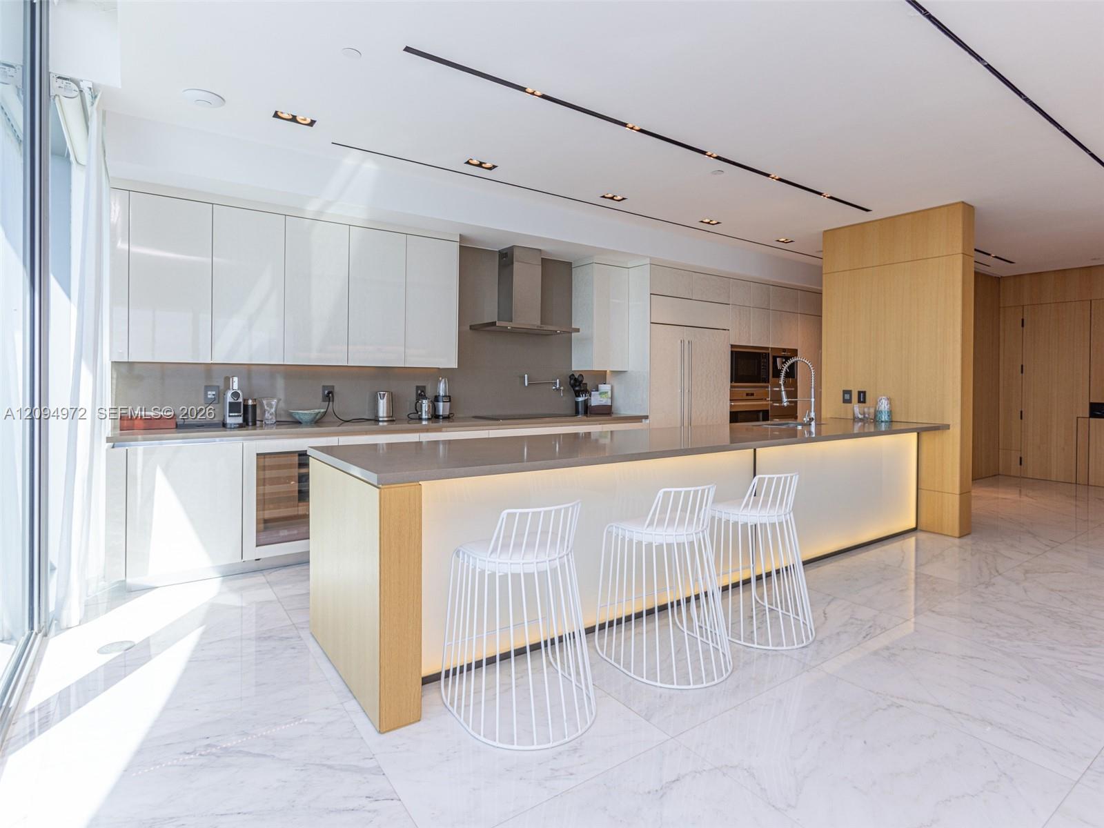 Modern kitchen with an illuminated island, three white stools, white cabinetry, and marble floors.