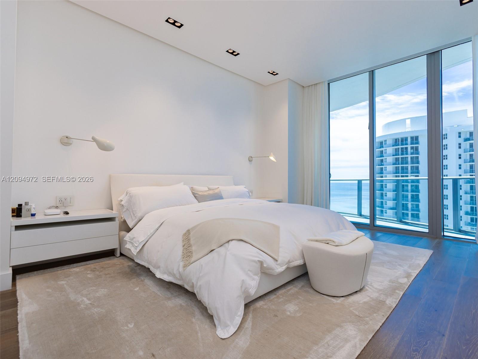 Furnished bedroom with a white bed and floor-to-ceiling windows overlooking the ocean.