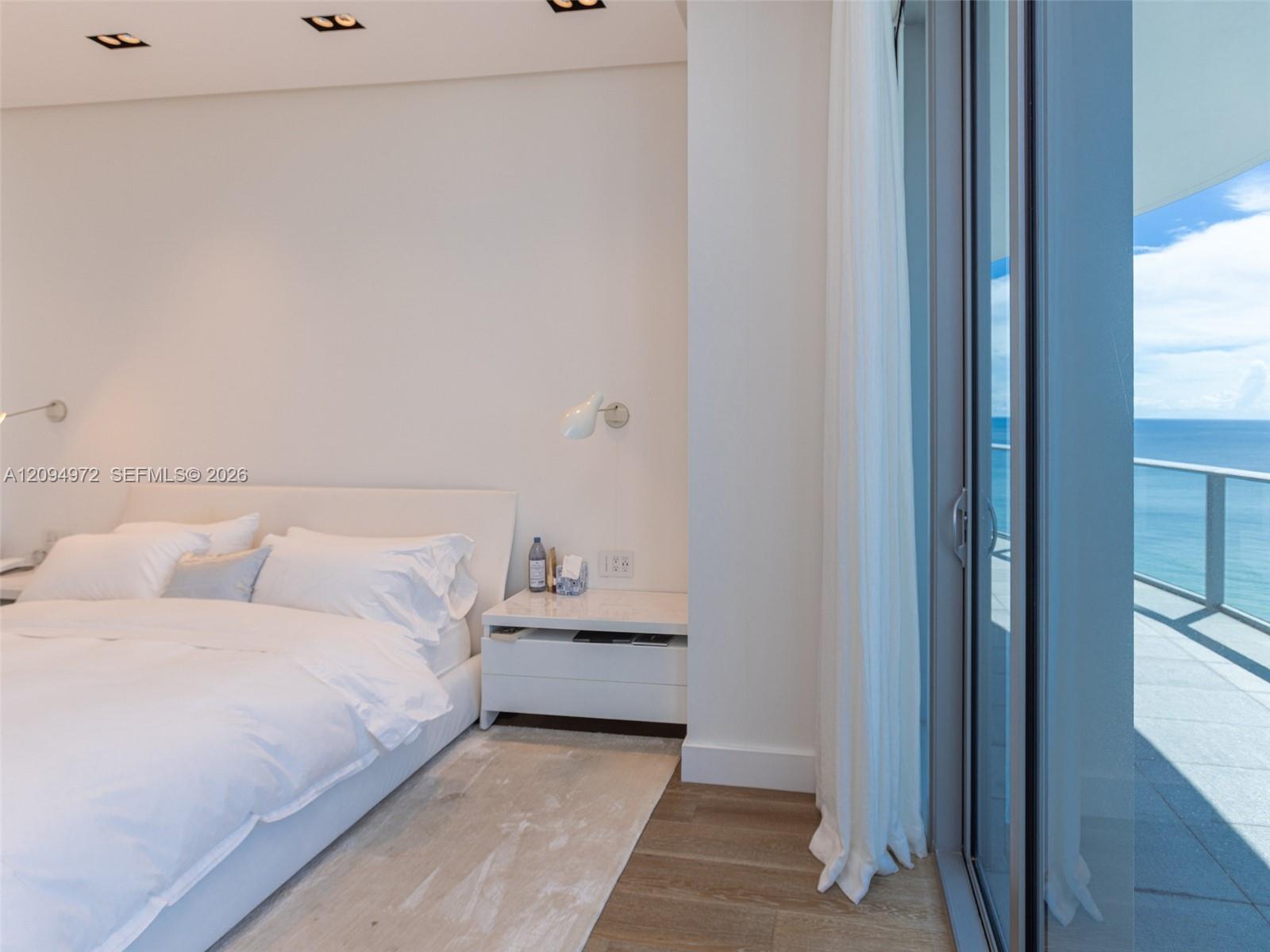 Bedroom with a white bed and floor-to-ceiling doors opening to a balcony overlooking the ocean.