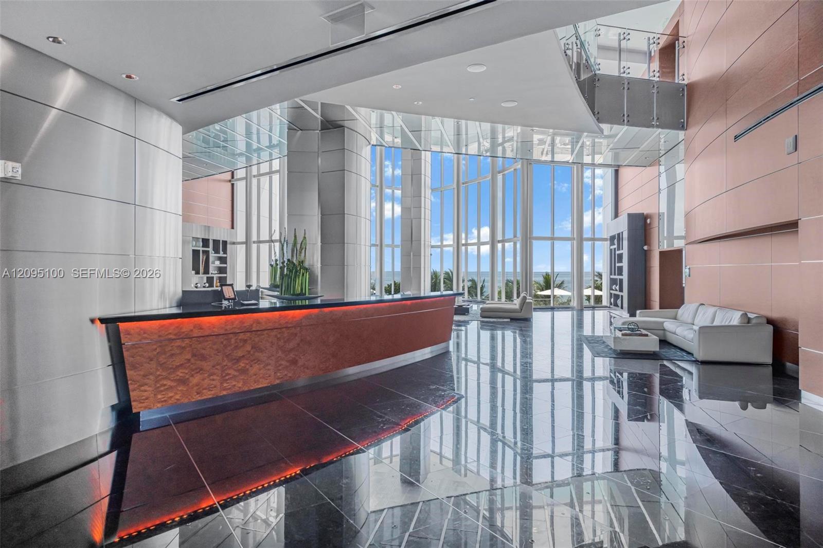 Condominium lobby with an illuminated reception desk, glossy floors, and ocean views through tall windows.