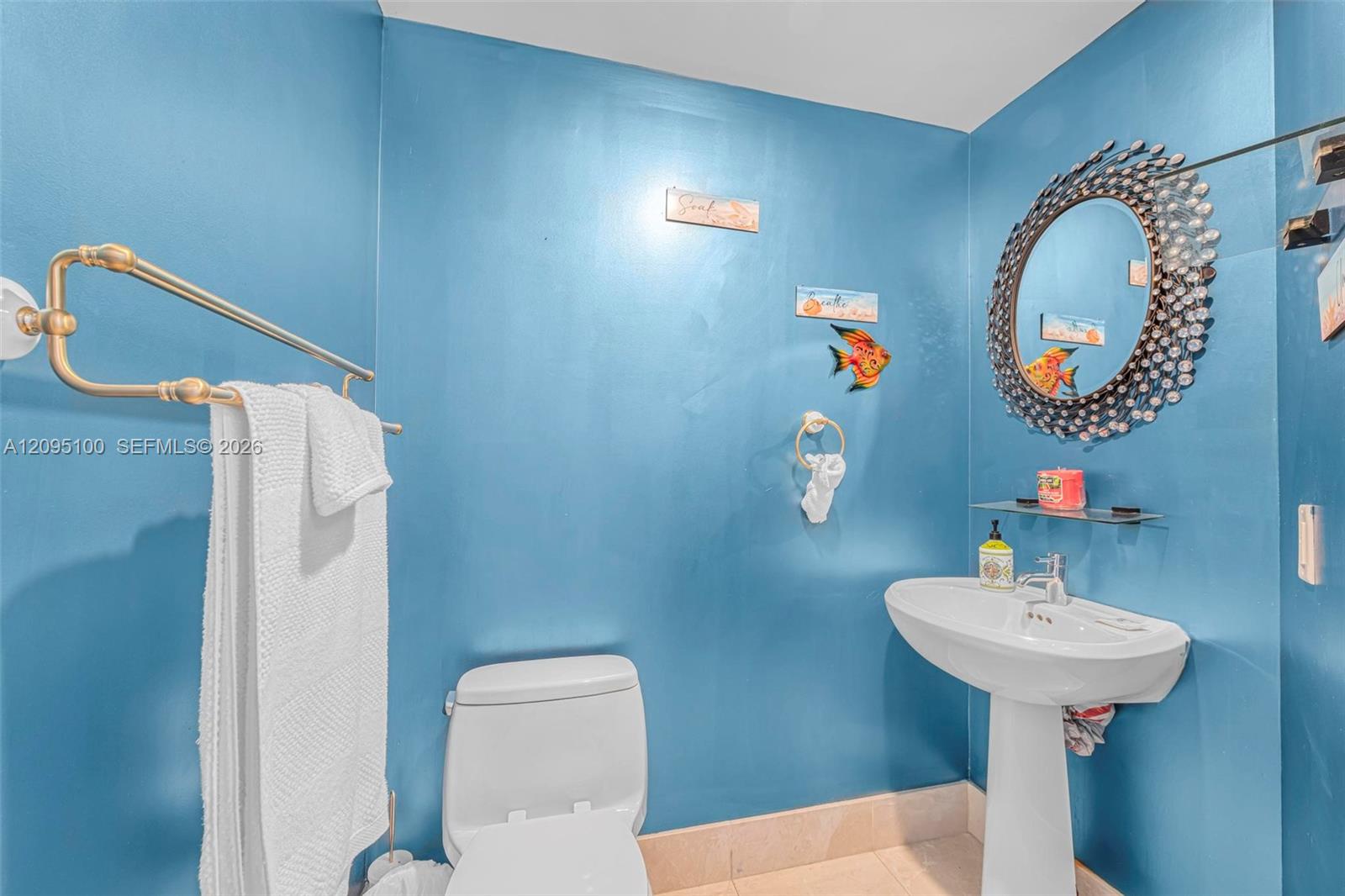 Blue bathroom with a white toilet, pedestal sink, ornate round mirror, and gold towel bar.