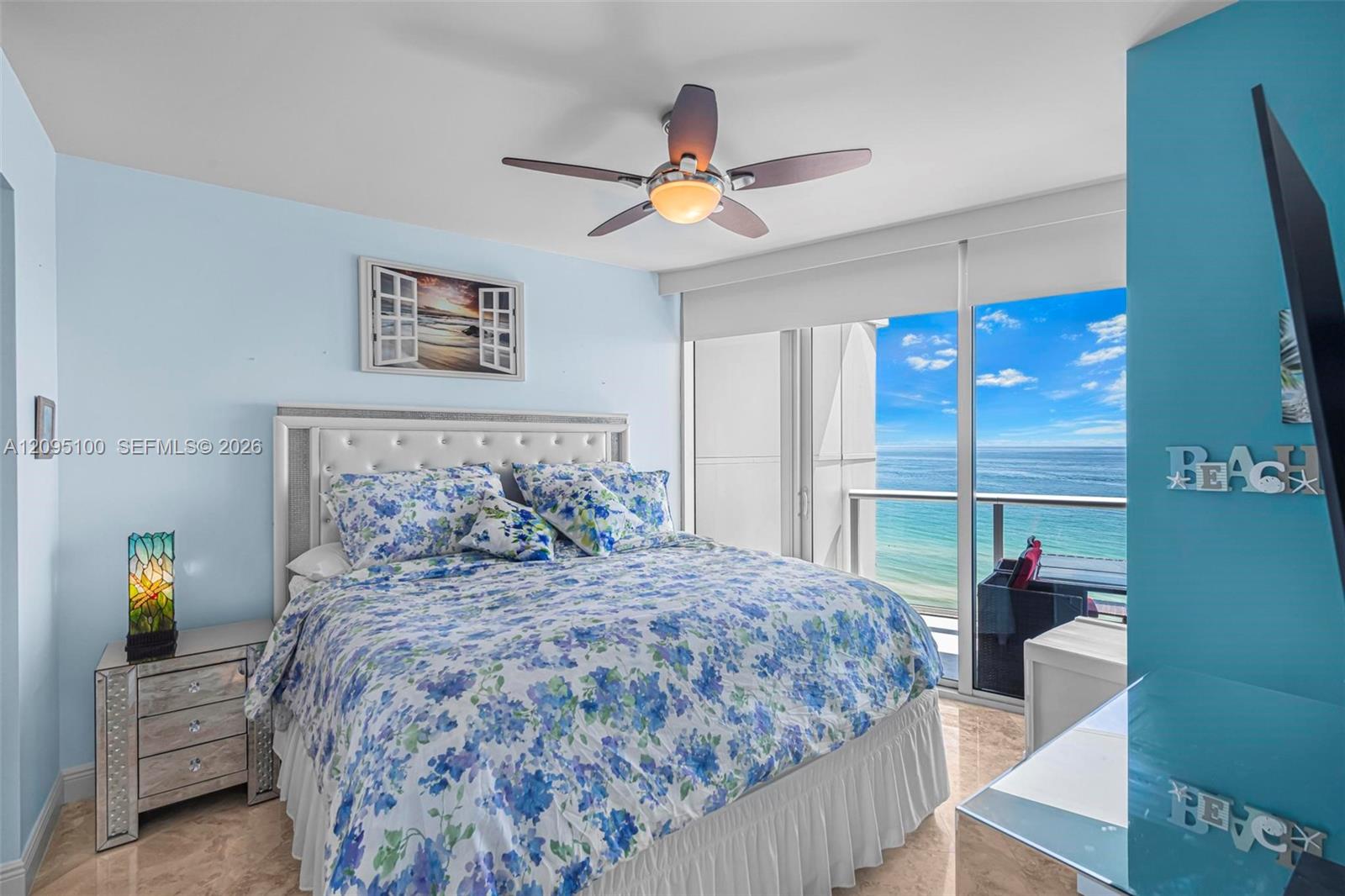 Ocean-facing bedroom with floral bedding, mirrored nightstand, and sliding doors to a balcony.
