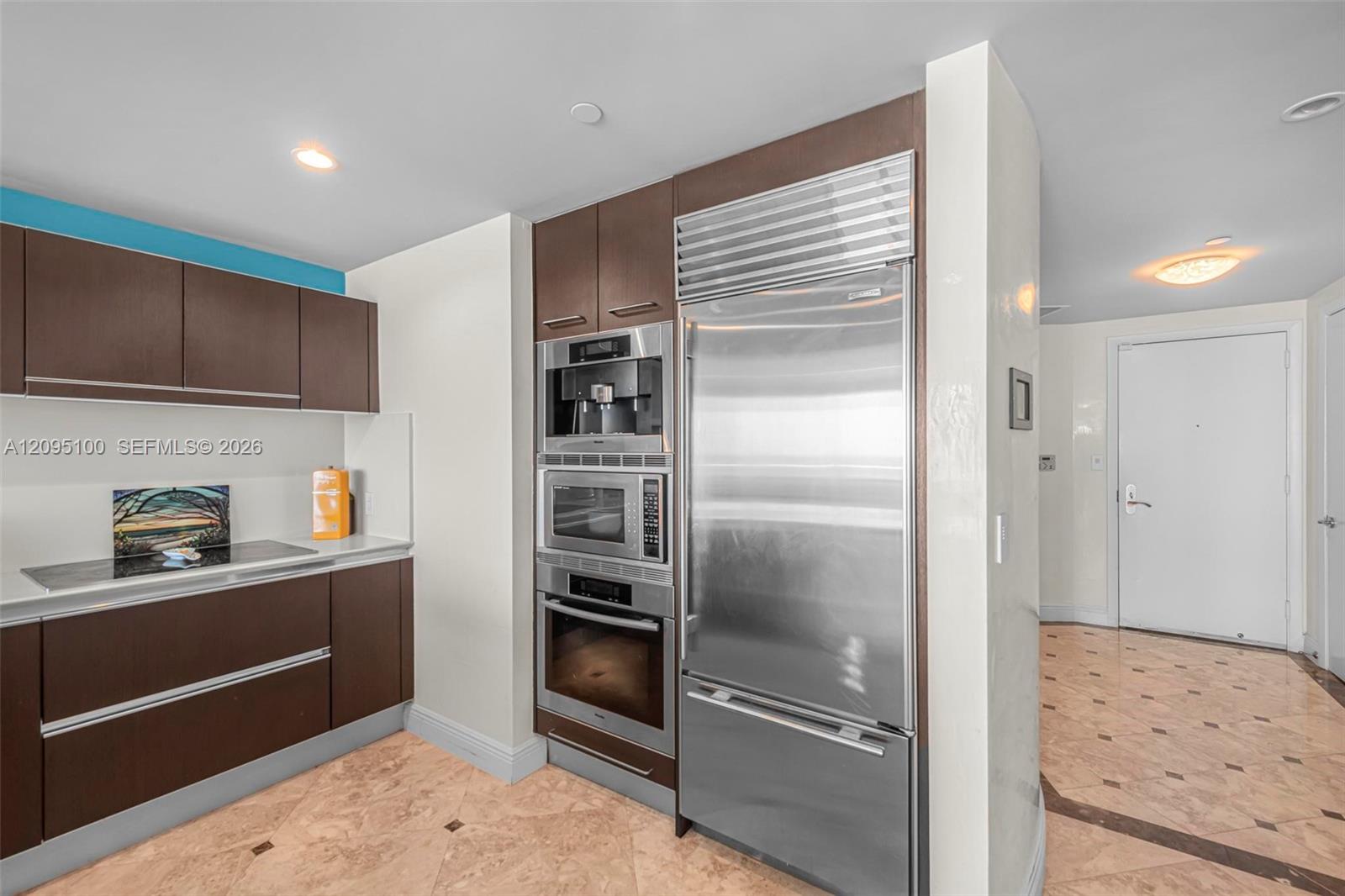 Condo kitchen with dark cabinetry, stainless steel appliances, and pale stone tile flooring.