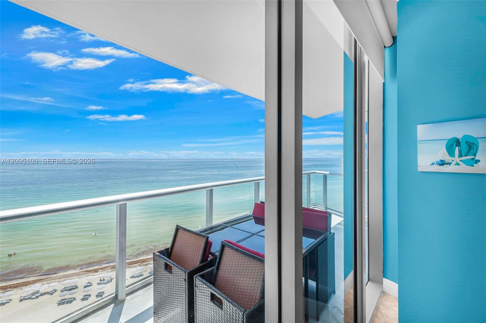 Oceanfront balcony with wicker chairs and glass railing overlooking the beach and sea.