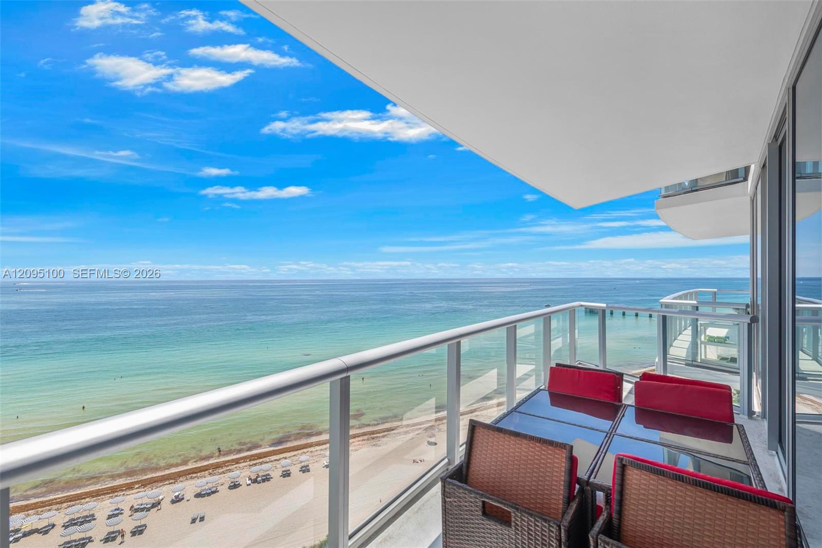 Furnished condo balcony overlooking turquoise ocean and sandy beach, with glass railings and wicker seating.