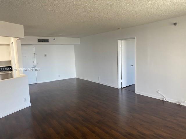 Empty living room with dark wood flooring, a kitchen pass-through, and an open doorway.