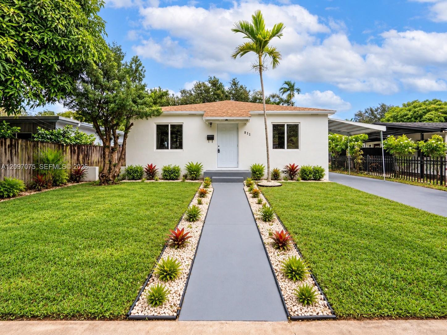 White single-story house with a landscaped front lawn, tropical plants, and a walkway to the entrance.