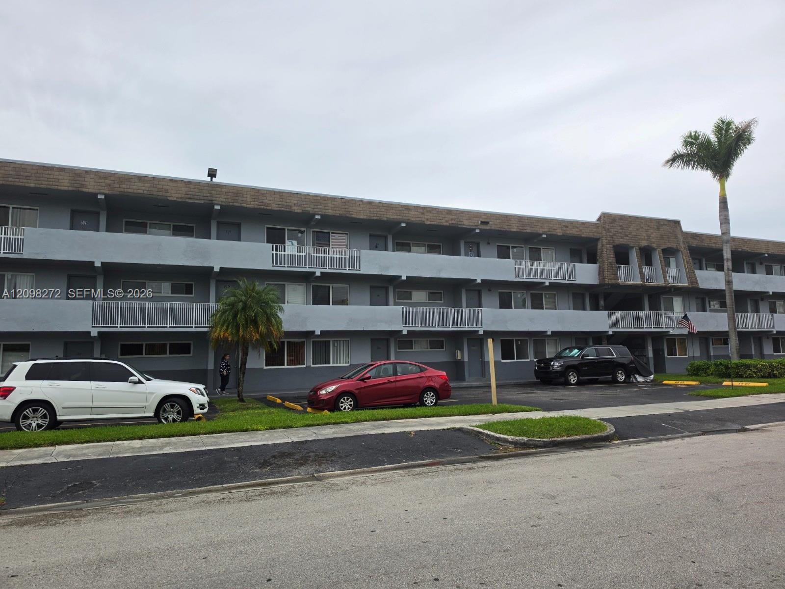 Three-story condominium building with long balconies, white railings and palm trees along the street.