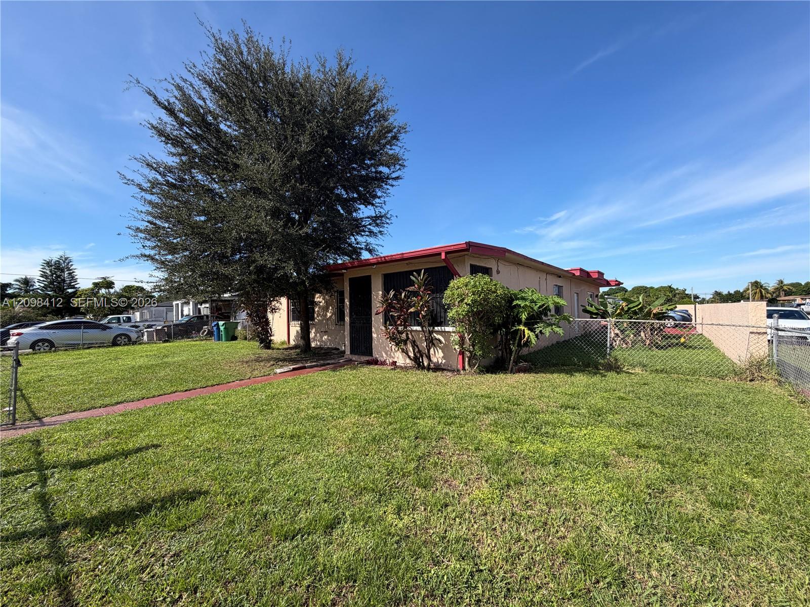 Single-story beige house with a red roof, lawn, chain-link fence, and large shade tree.
