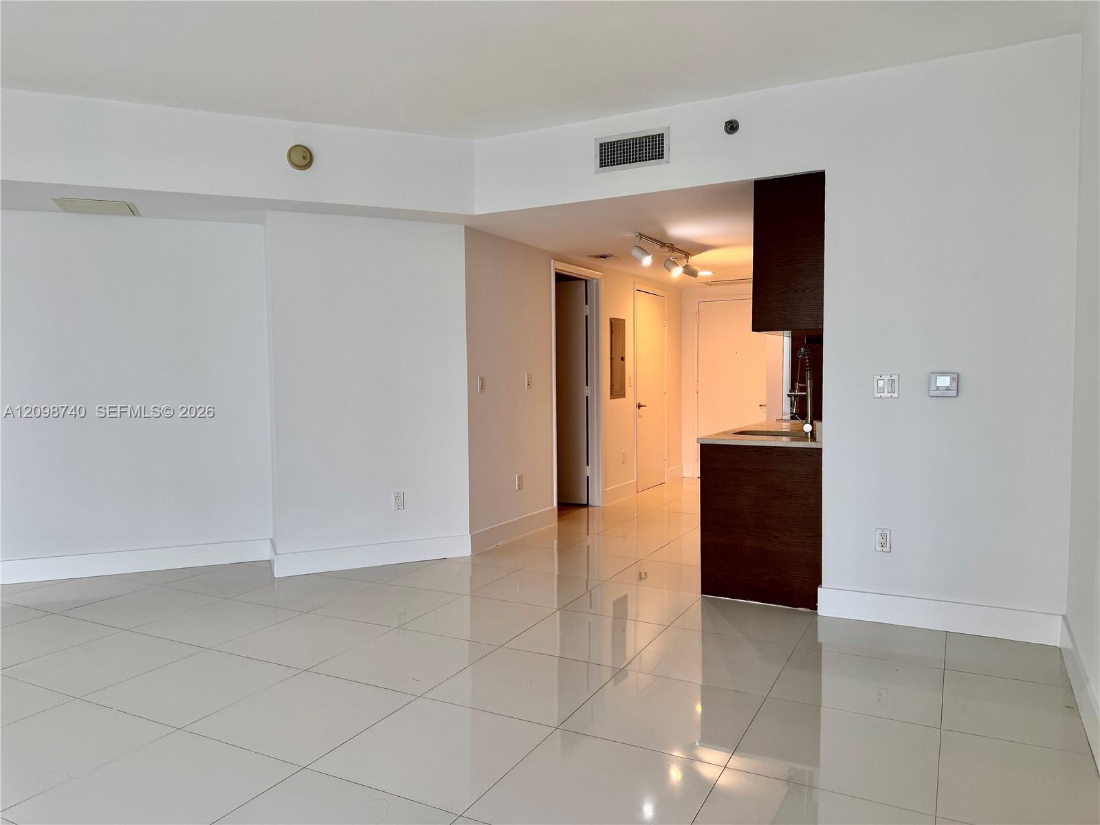 Empty condo living area with glossy white tile floors and a dark wood kitchen peninsula.