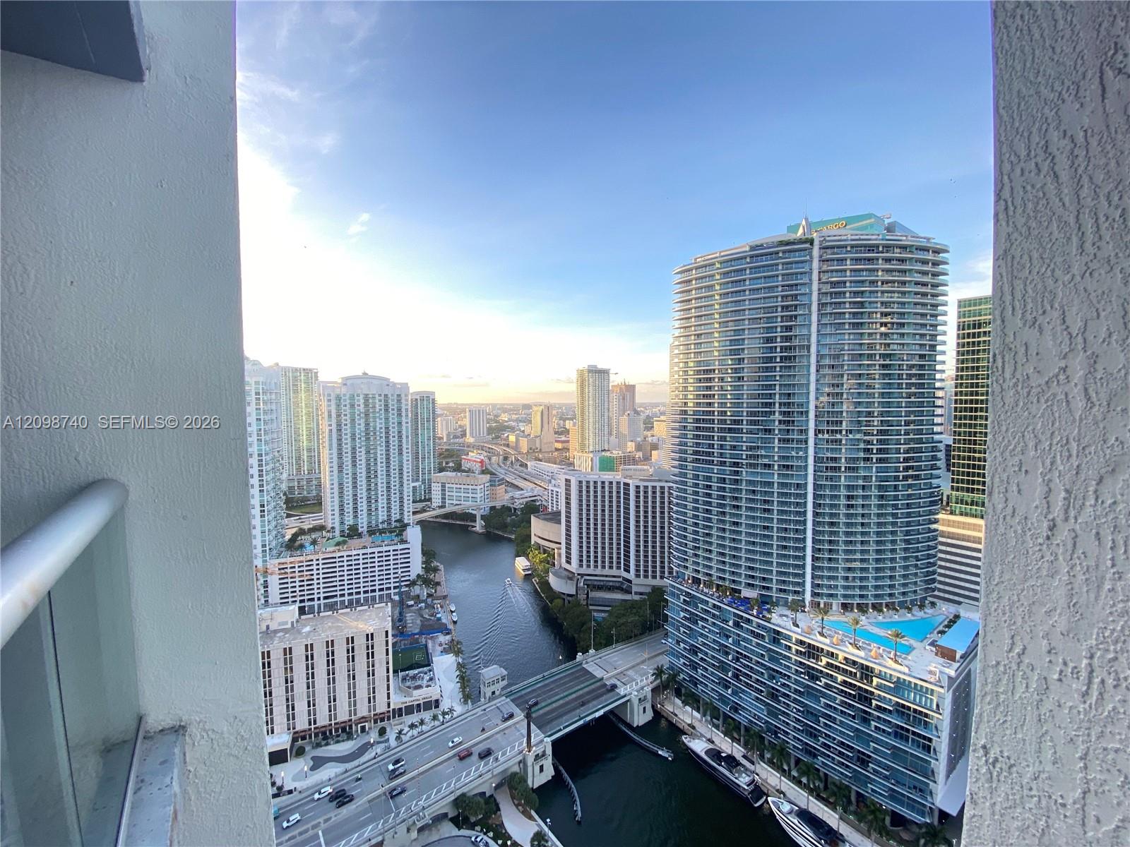 Miami skyline and canal framed by a balcony, with high-rise towers and bridges in warm evening light.