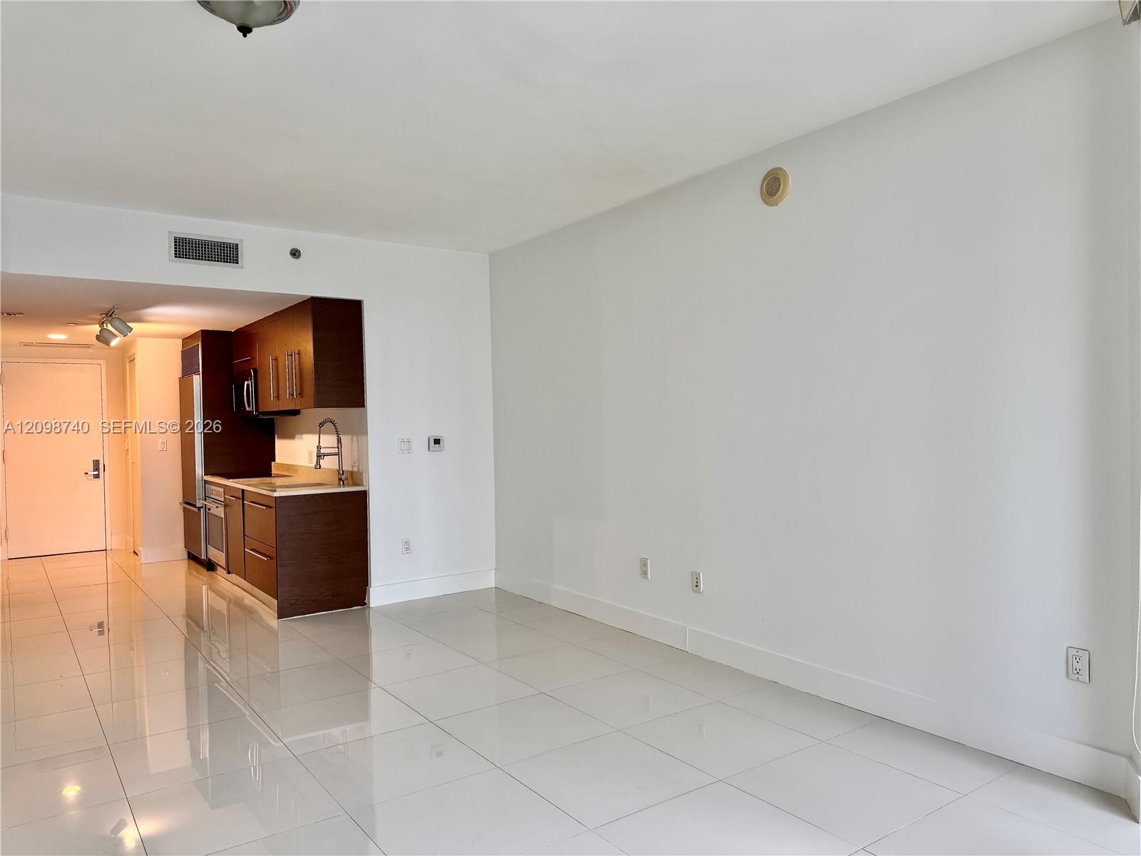 Vacant condo living area with glossy tile floors and a compact dark-wood kitchen.