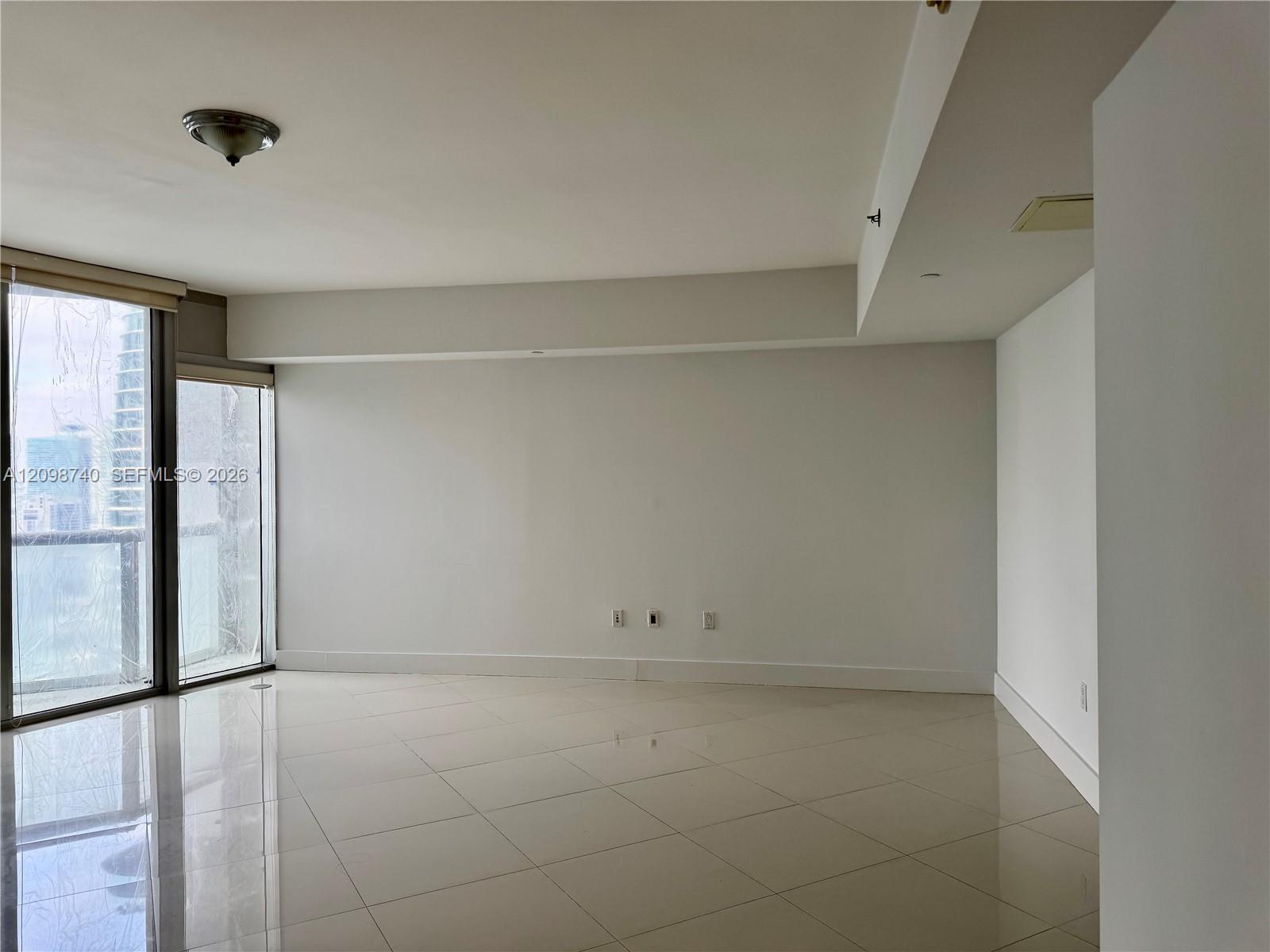Empty tiled living room with sliding balcony doors and neighboring buildings visible outside.