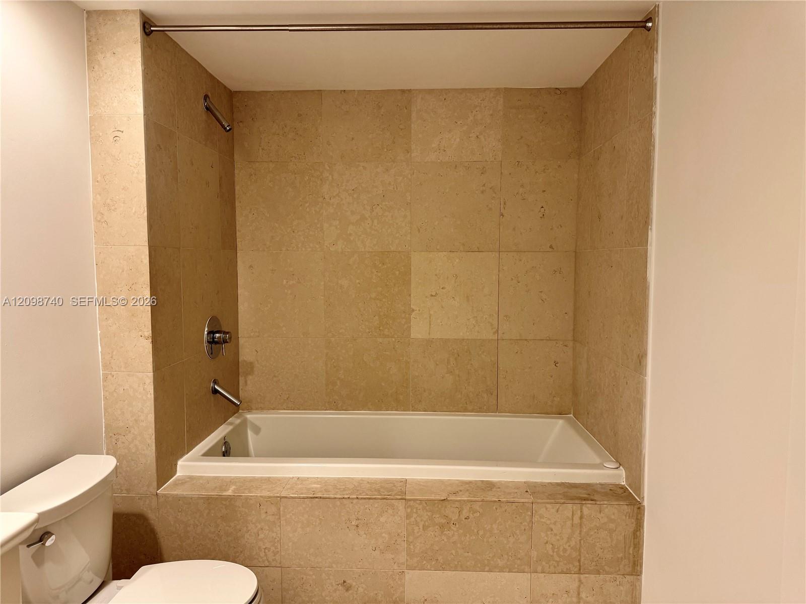 Bathroom bathtub in a beige stone-tile alcove with chrome shower fixtures and a partially visible toilet.