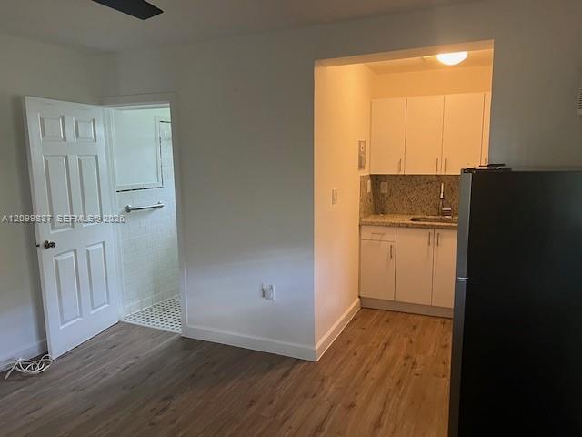 Vacant room with a compact granite-counter kitchen, black refrigerator, wood-look floors, and bathroom doorway.