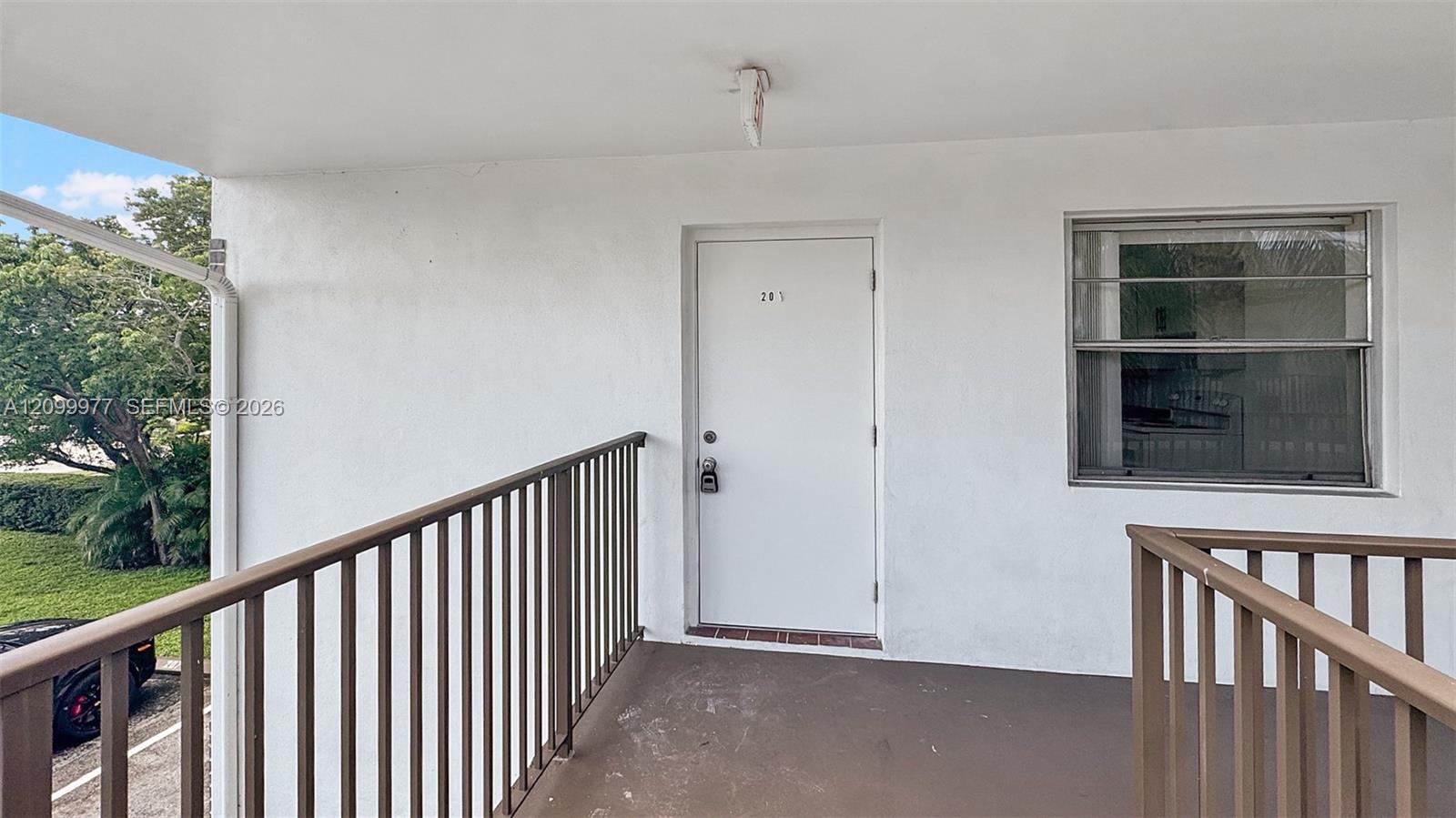 Covered condo entry balcony with a white door, window, and brown railings overlooking trees and parking.