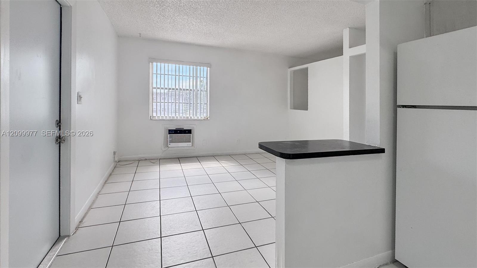 Vacant studio interior with tiled floors, a window air conditioner, breakfast counter, and refrigerator.