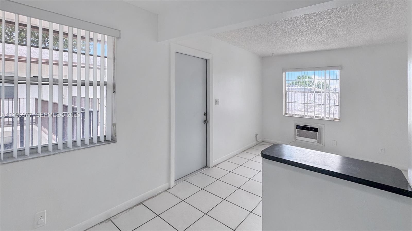 Empty tiled interior with a dark countertop peninsula, vertical blinds, and a window air conditioner.