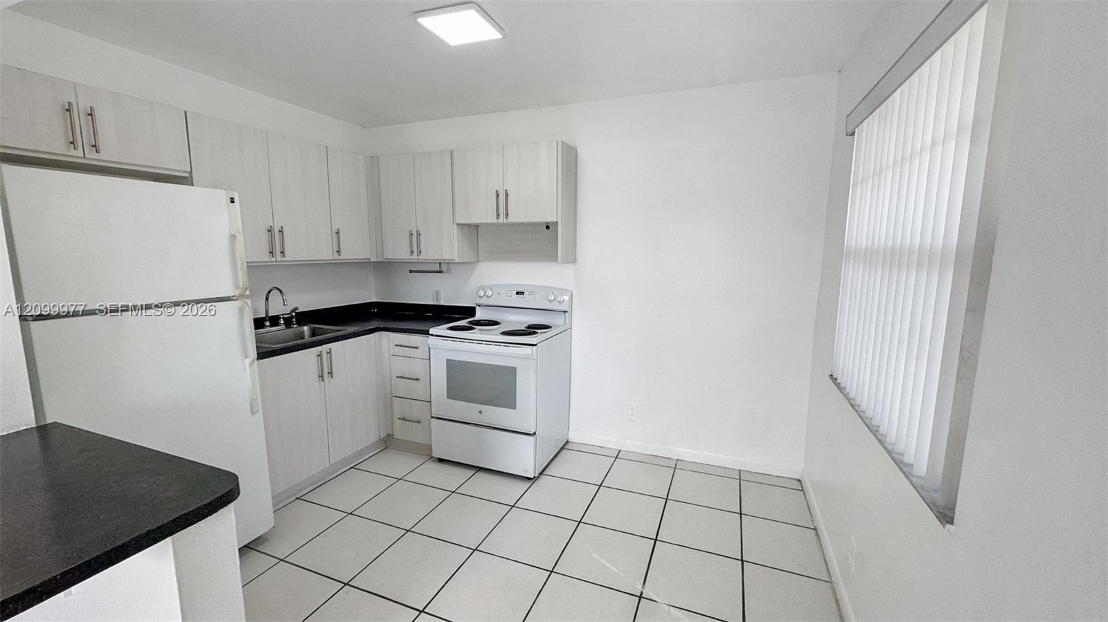 Vacant kitchen with white cabinets, black countertops, white appliances, and a large window with vertical blinds.