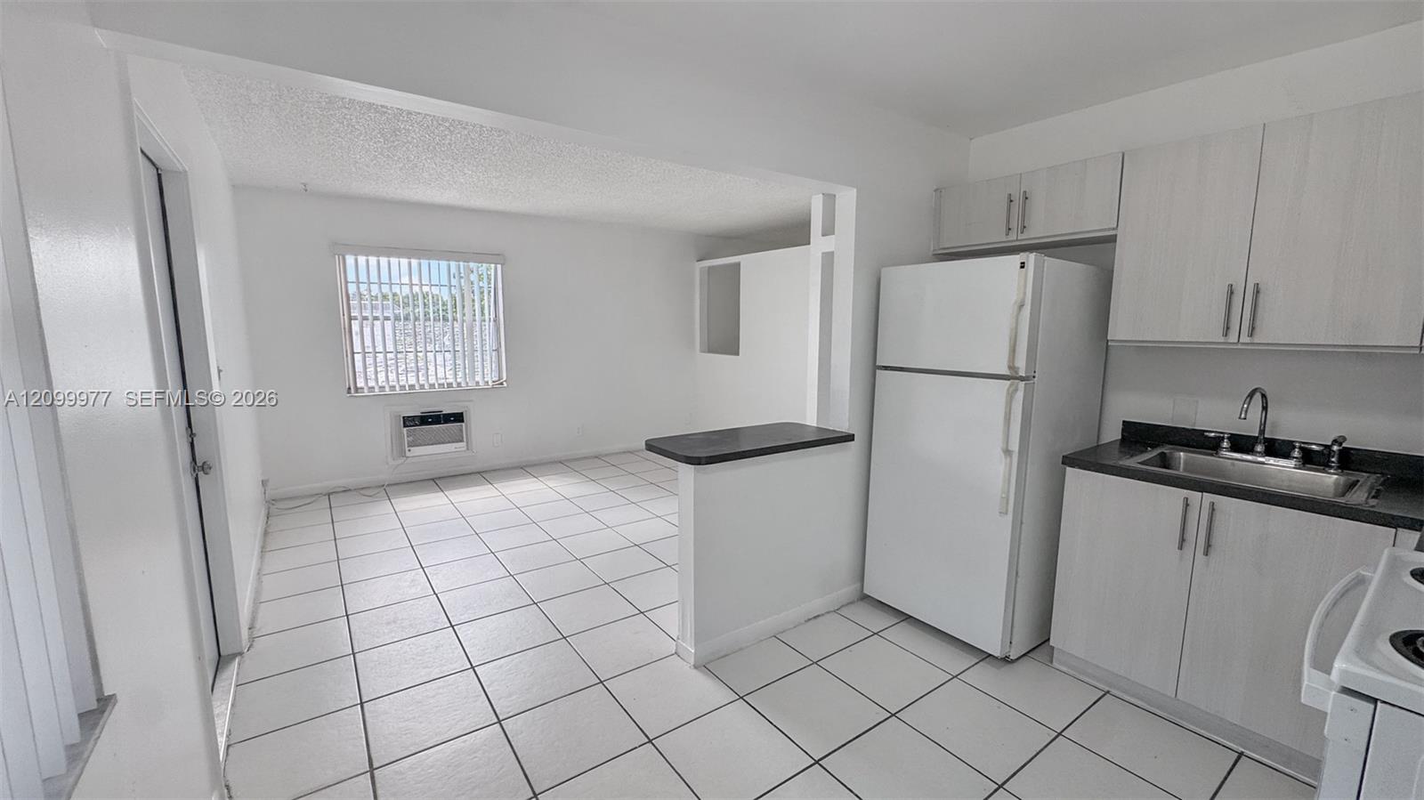 Vacant kitchen opening to a tiled living area, with white cabinets and a black breakfast counter.