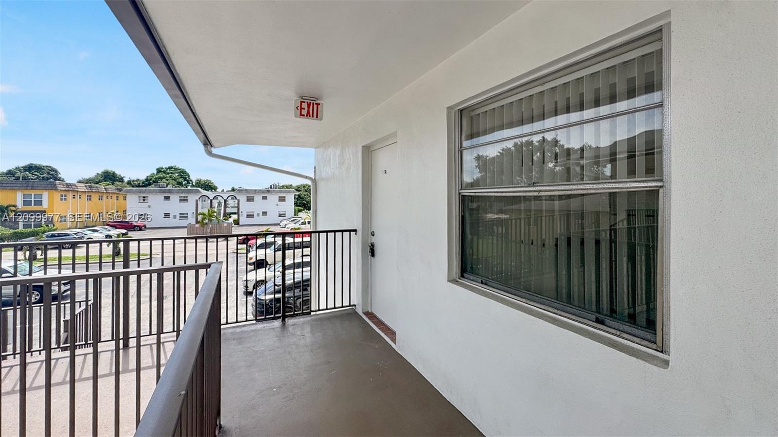 Covered apartment balcony with black railing overlooking a parking lot and neighboring buildings.